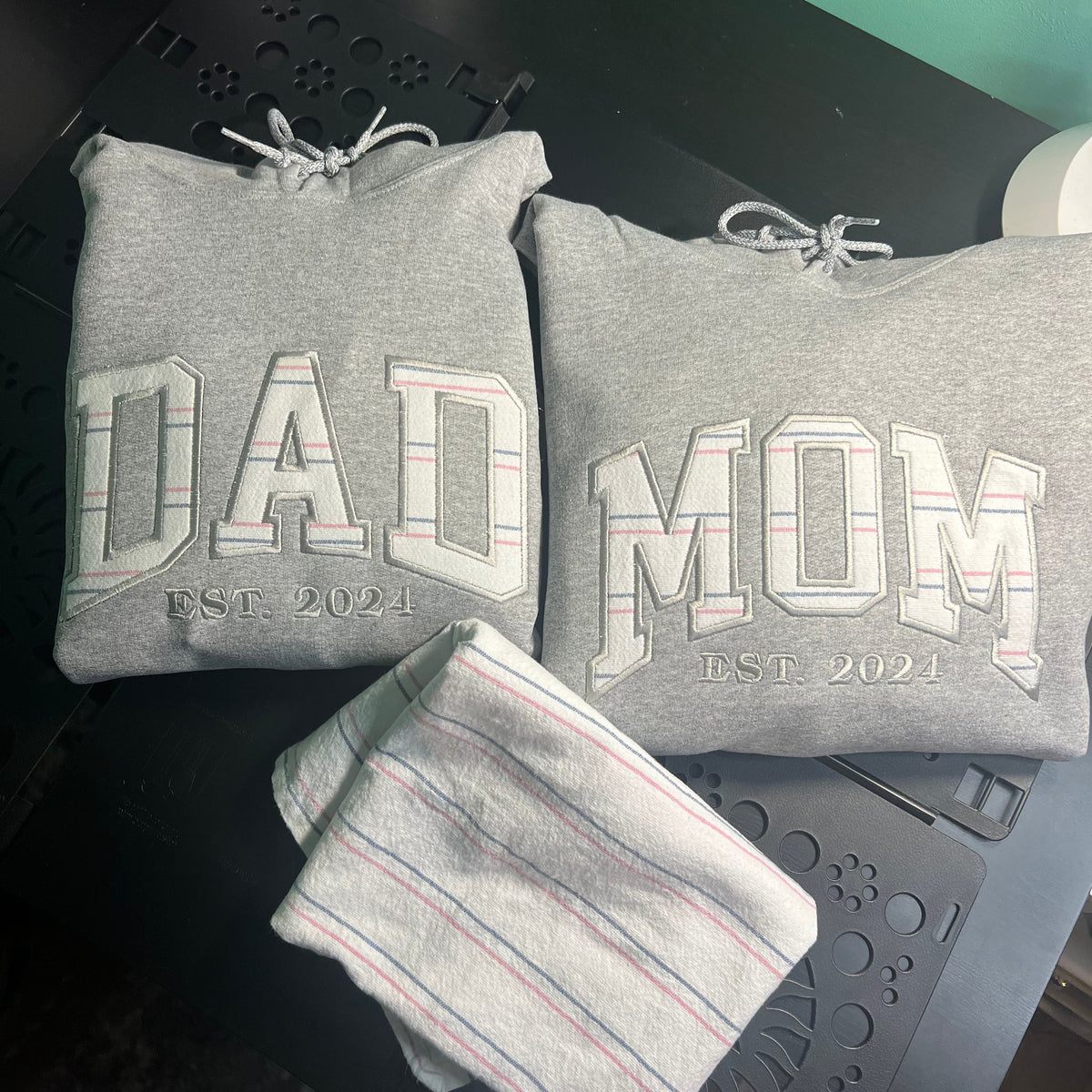 Two folded heather gray drawstring hoodies with 'DAD' and 'MOM' text on the front, plus a matching striped towel, laid on a dark surface.