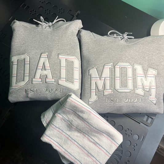 A folded heather gray sweatshirt with 'DAD' and 'MOM' printed in large outlined letters, plus 'EST. 2024' below each word, shown alongside a matching striped towel on a dark surface.